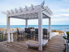 Um deck de madeira com pérgola, cadeiras e um banco com vista para o oceano.