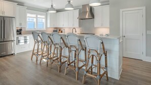 A modern kitchen with white cabinetry, a stainless steel refrigerator, a built-in oven, and a large island with bar stools. - Love Kure by Carolina Retreats (Kure Beach)