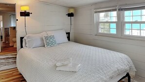 A bedroom with a large bed, two bedside lamps, a window with a view, and wooden flooring. - Toes in the Sand by Carolina Retreats (Carolina Beach)