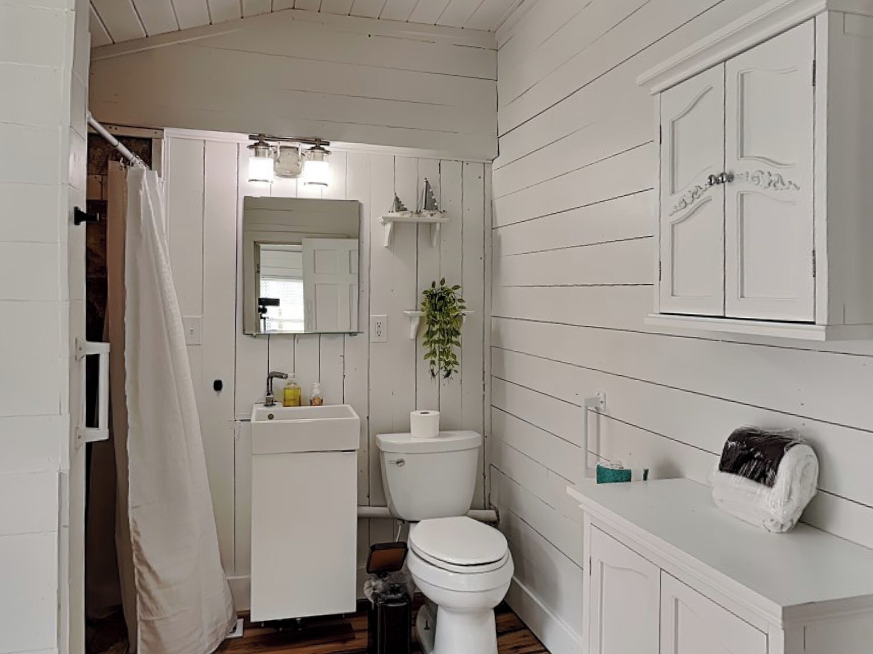 A small, white, wooden-paneled bathroom with a toilet, sink, and a mirror above the sink. There is a shower curtain partially open, and a small plant on the wall.