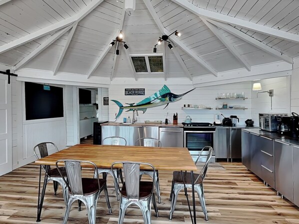 A modern kitchen with a wooden dining table, metal chairs, and a large fish decoration. - Toes in the Sand by Carolina Retreats (Carolina Beach)