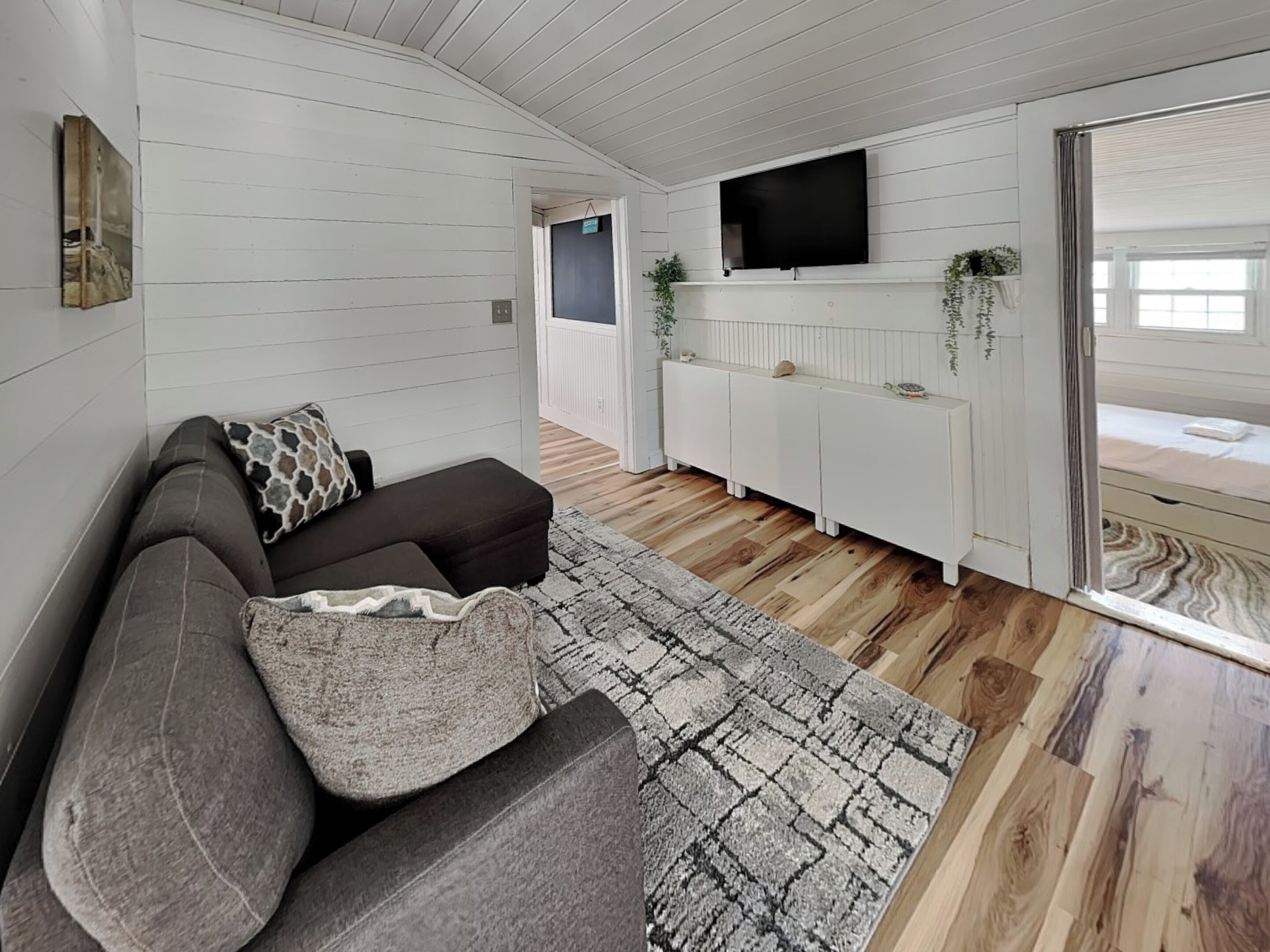 A modern living room with a grey sofa, patterned rug, wooden flooring, white walls, a mounted TV, and a sliding glass door leading to a bedroom.