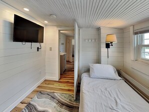 A bedroom with a bed, a TV mounted on the wall, a lamp, and a window. - Toes in the Sand by Carolina Retreats (Carolina Beach)