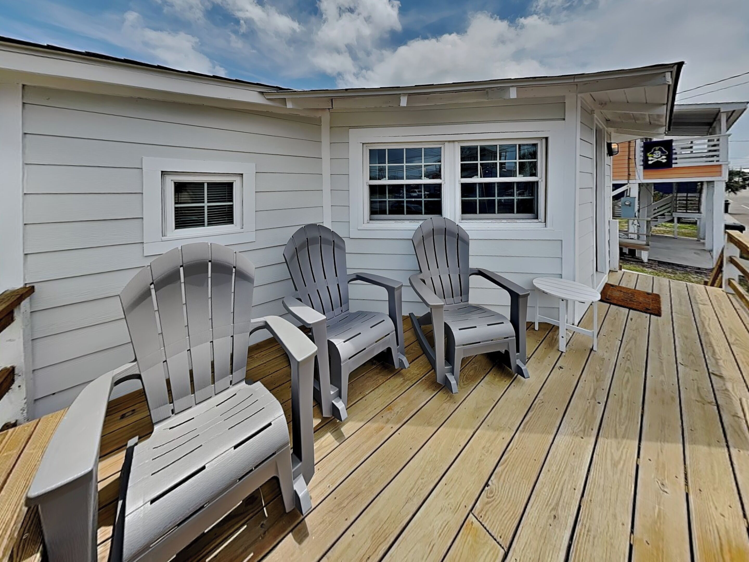 A deck with Adirondack chairs and a small table, wooden deck flooring, and a white building with windows.