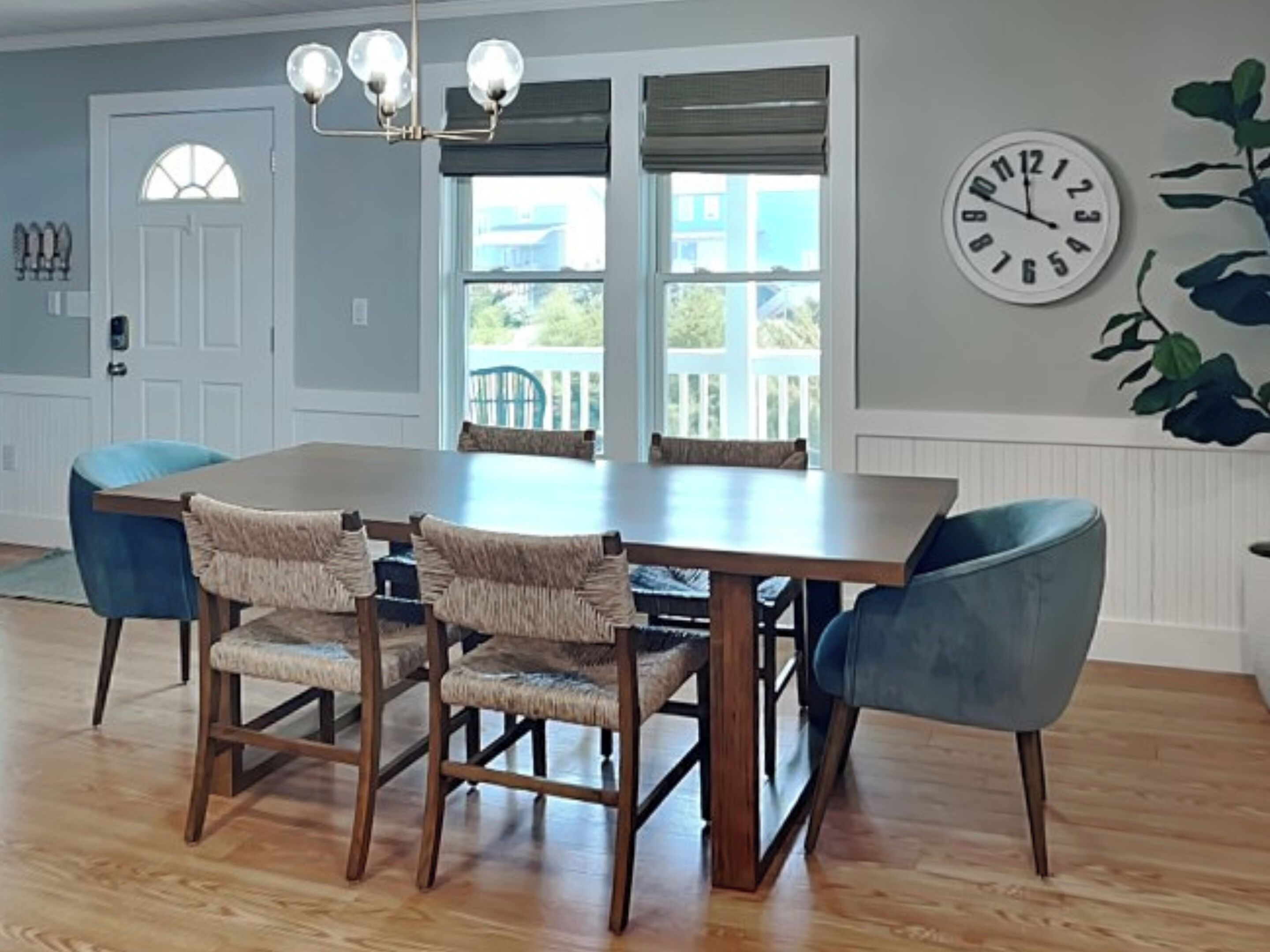 A dining room with a wooden table and chairs, a chandelier, a clock, and a view of the outside.