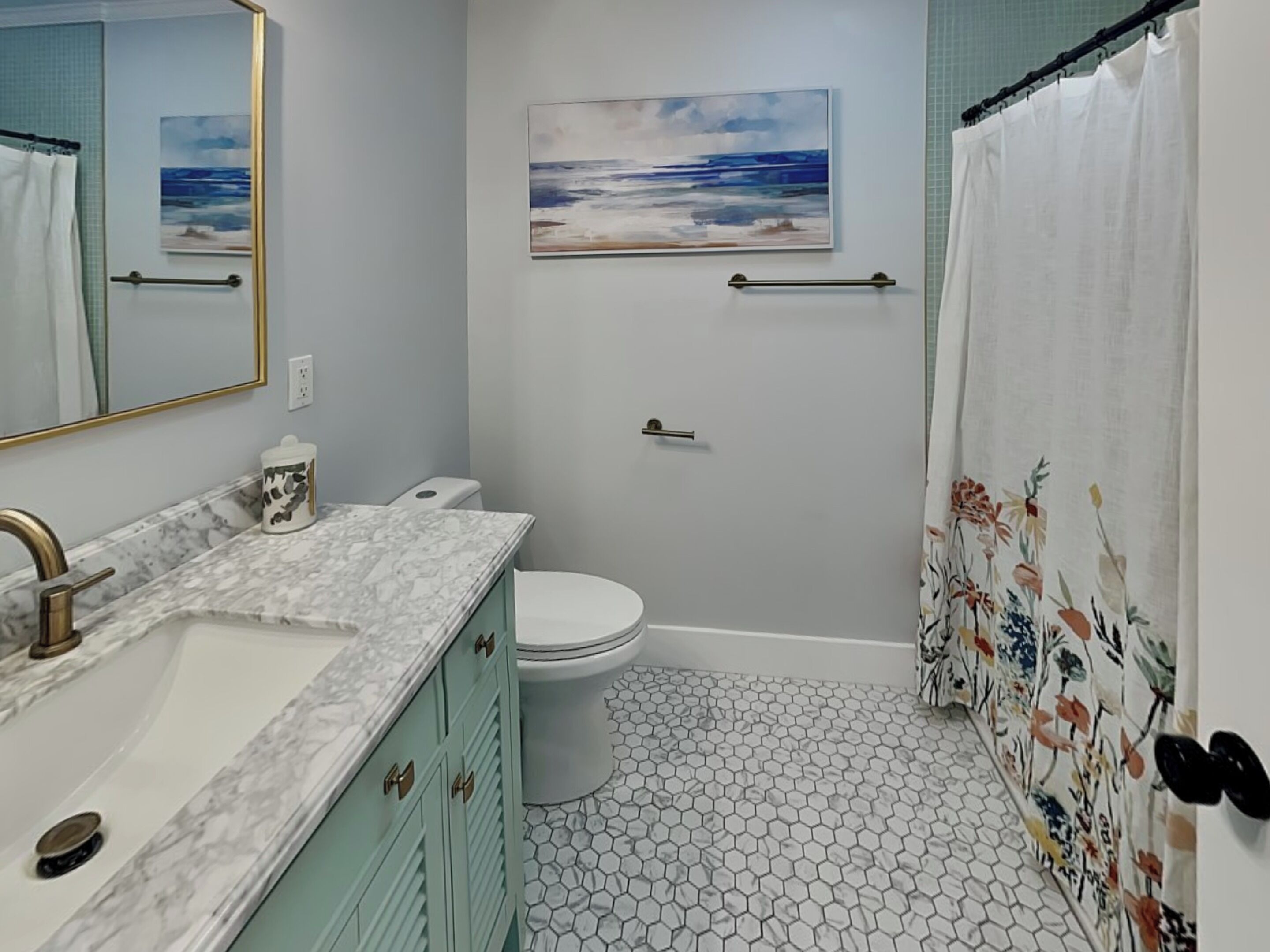 Ein Badezimmer mit Marmorwaschbecken, Toilette, Dusche mit floralem Vorhang, einem Strandgemälde und sechseckiger Fliesenverkleidung.