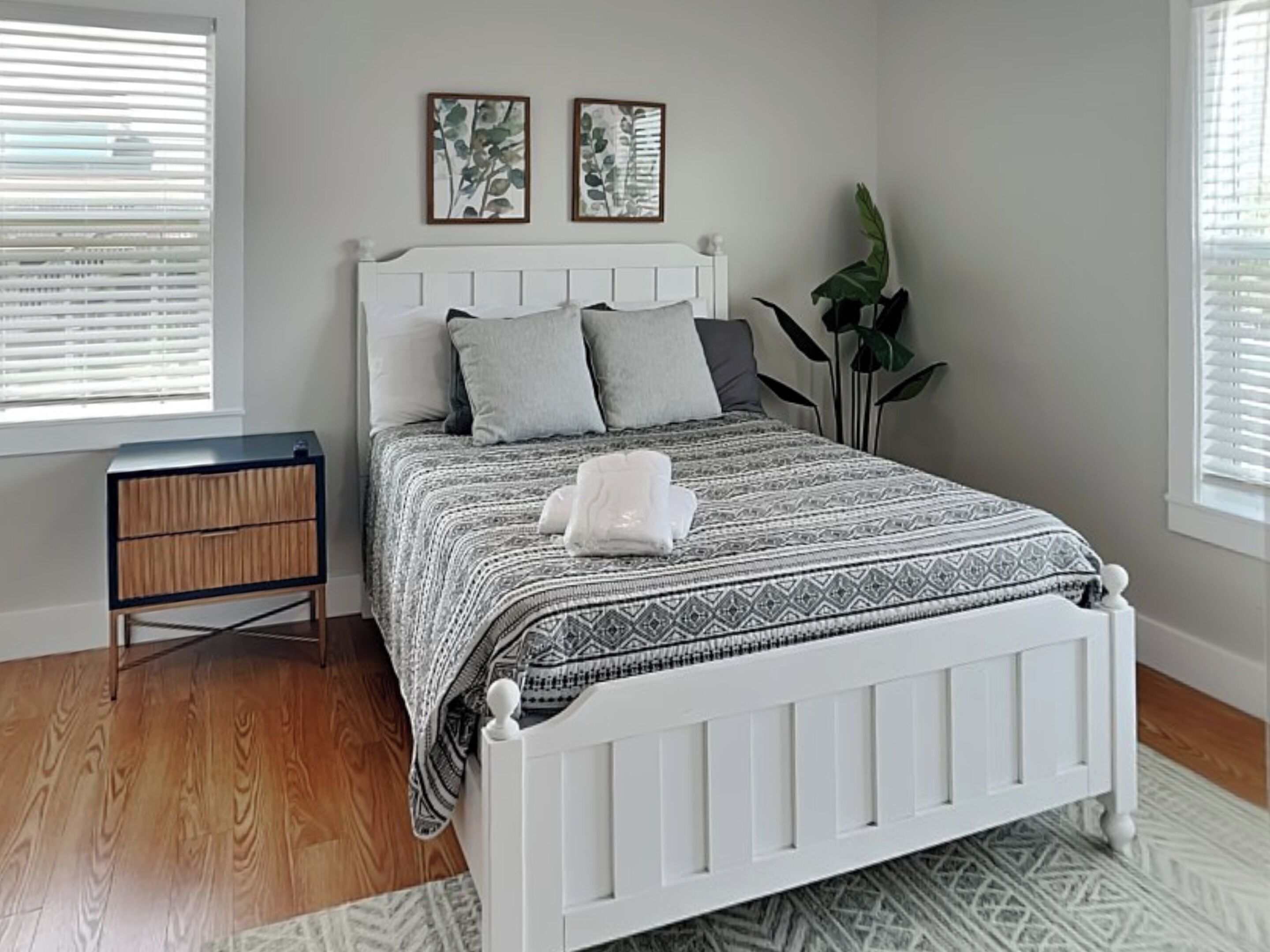 Un dormitorio con cama blanca, ropa de cama con estampado gris y blanco, una mesita de noche y un gran ventanal con persianas.