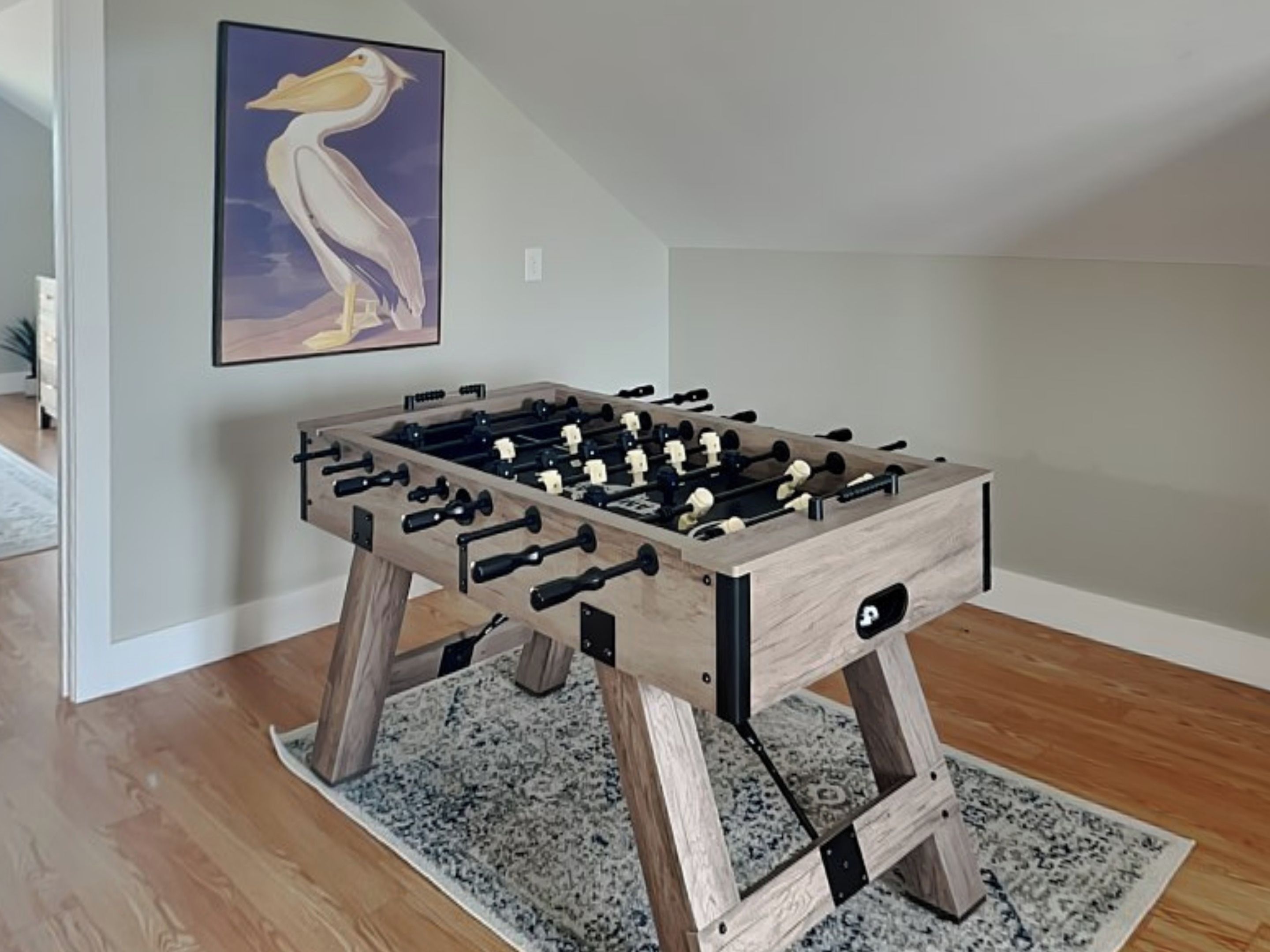 Une table de baby-foot en bois se trouve dans une pièce dotée d’un tapis gris et d’une œuvre d’art encadrée accrochée au mur.