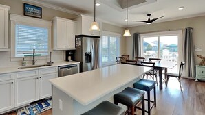A modern kitchen with white cabinets, a large island with bar stools, a refrigerator, and a dining area with a table and chairs. - Sunset Nonsense by Topsail Realty (Holly Ridge)