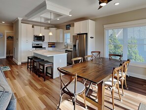 A modern kitchen with white cabinetry, a central island, and a dining area with wooden chairs. - Sunset Nonsense by Topsail Realty (Holly Ridge)