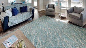 A living room with a blue patterned rug, a grey sofa with blue cushions, a grey armchair, and a small round table.