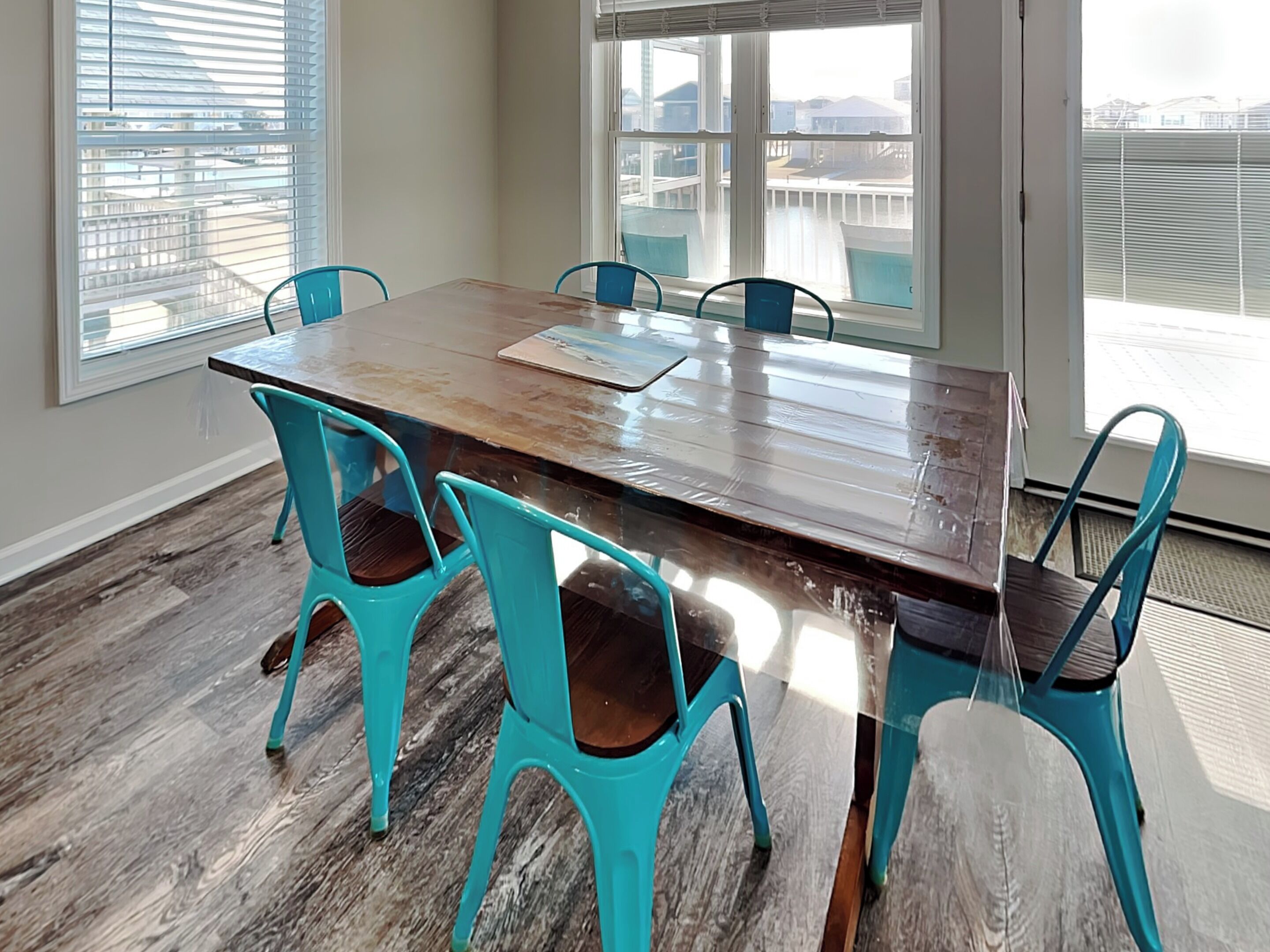 A dining area with a wooden table and turquoise chairs, large windows with blinds, and a view of a balcony.