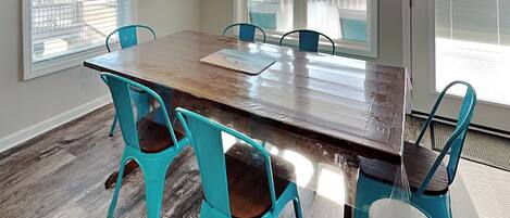 A dining area with a wooden table and turquoise chairs, large windows with blinds, and a view of a balcony.