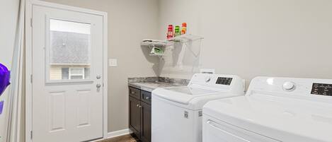 A laundry room with two white front-loading washers and dryers, a wooden cabinet, and a shelf with laundry supplies.