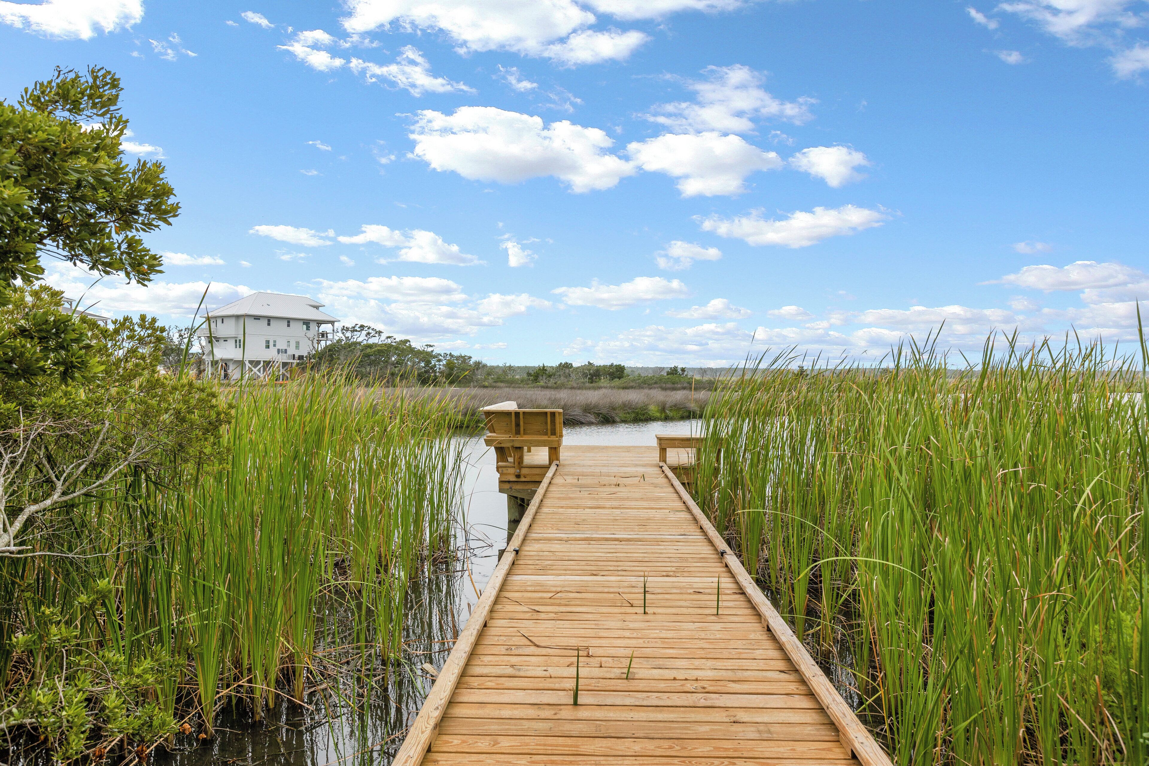 Une jetée en bois qui mène à une maison entourée de hautes herbes et bordée par l’eau.
