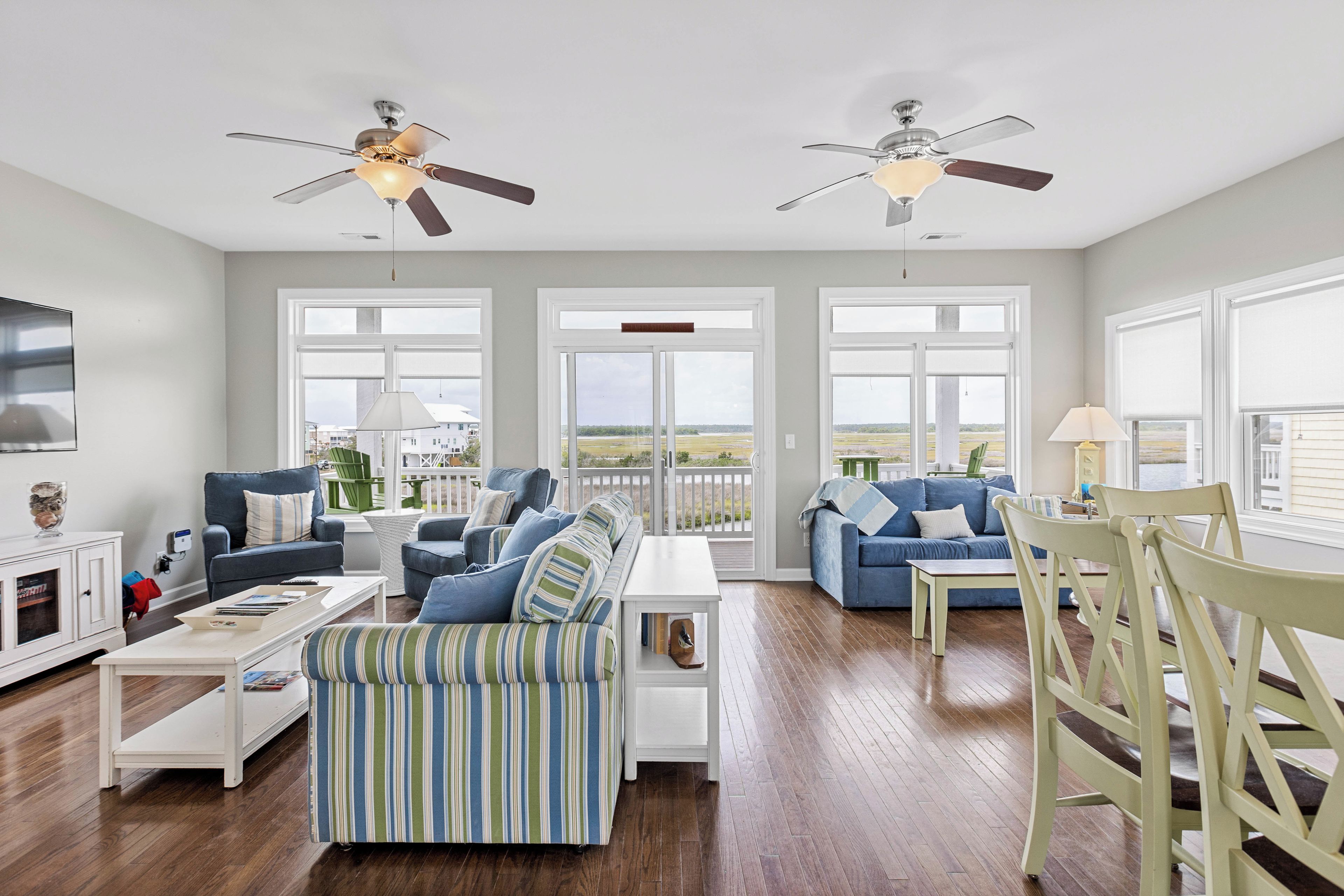 A spacious living room with two sofas, a coffee table, and a dining table. There are ceiling fans, large windows, and a view of the outdoors.