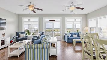 A spacious living room with two sofas, a coffee table, and a dining table. There are ceiling fans, large windows, and a view of the outdoors.