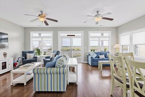 A spacious living room with two sofas, a coffee table, and a dining table. There are ceiling fans, large windows, and a view of the outdoors. - Palm Beach North by Topsail Realty (Sneads Ferry)