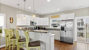 A kitchen with white cabinets, a stainless steel refrigerator, a microwave, and a bar area with stools. - Palm Beach North by Topsail Realty (Sneads Ferry)