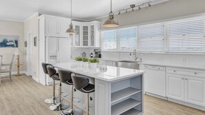 A modern kitchen with white cabinetry, a large island with bar stools, pendant lighting, and a view of the outdoors through large windows. - Topsail Escape by Topsail Realty (Sneads Ferry)