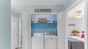 A laundry room with a 'Welcome to the Beach' sign, a washing machine, dryer, and shelves with laundry supplies.
