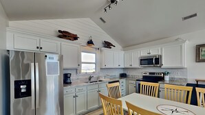 Une cuisine moderne avec des meubles blancs, des appareils en acier inoxydable, un coin repas avec des chaises en bois et une grande fenêtre qui laisse entrer la lumière naturelle.