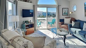 A spacious living room with a large TV, comfortable seating, a balcony with chairs, and a ceiling fan.