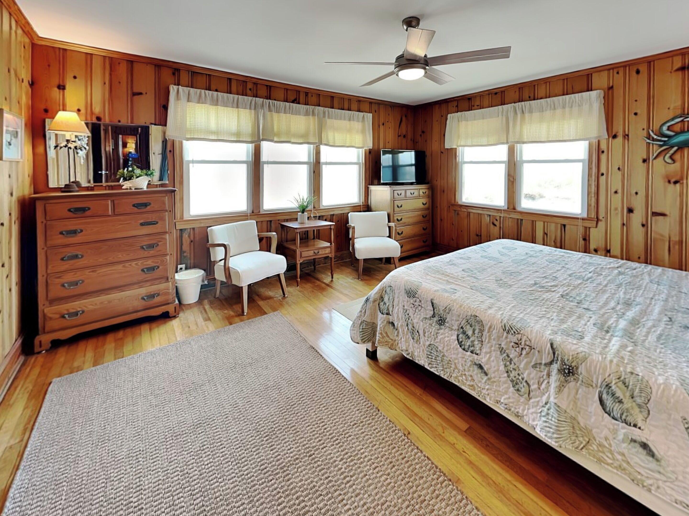 A wooden-paneled room with a bed, a desk, a chair, a dresser, and a ceiling fan.