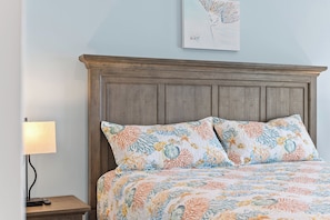 A bedroom with a wooden headboard, a bed with floral-patterned bedding, a nightstand with a lamp, and a wall-mounted artwork. - Seadragon 1 by Carolina Retreats (Carolina Beach)
