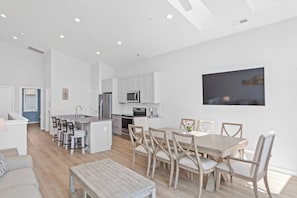 A modern kitchen with white cabinets, a dining area with a wooden table and chairs, and a large window. - Seadragon 1 by Carolina Retreats (Carolina Beach)