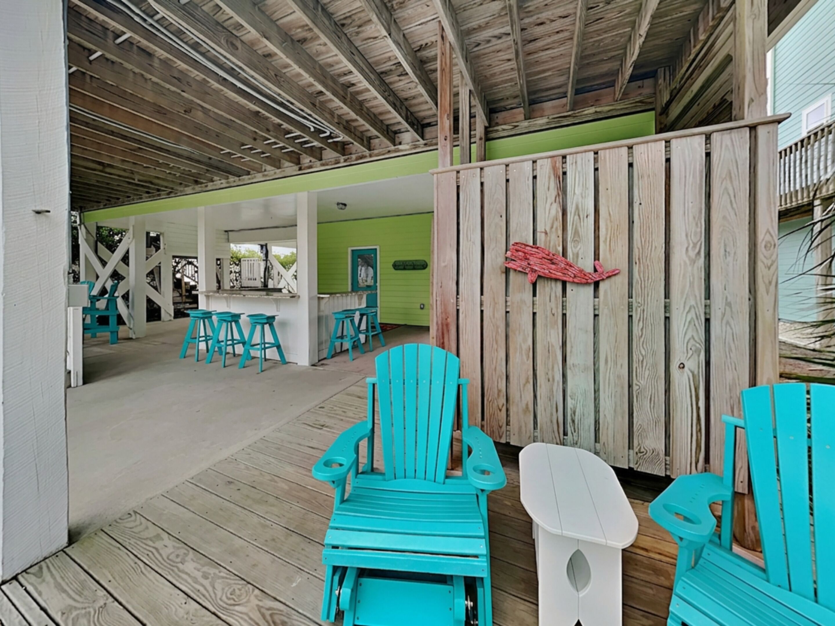 A covered outdoor area with wooden flooring, turquoise Adirondack chairs, and a white Adirondack chair.