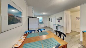 A compact kitchen with white cabinets, a refrigerator, and a dining area with a wooden table and blue chairs. - Catch A Wave by Topsail Realty (Holly Ridge)