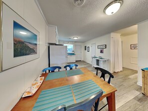 A compact kitchen with white cabinets, a refrigerator, and a dining area with a wooden table and blue chairs. - Catch A Wave by Topsail Realty (Holly Ridge)