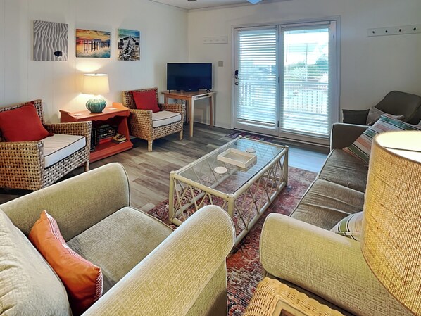 A living room with wicker furniture, a glass coffee table, a flat-screen TV, and a large sliding glass door to the outdoors. - Catch A Wave by Topsail Realty (Holly Ridge)