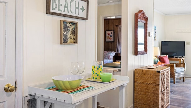 A room with a white wall, a framed 'BEACH' sign, a painting, a wicker side table with a bowl, glasses, and a checkered napkin holder. - Catch A Wave by Topsail Realty (Holly Ridge)