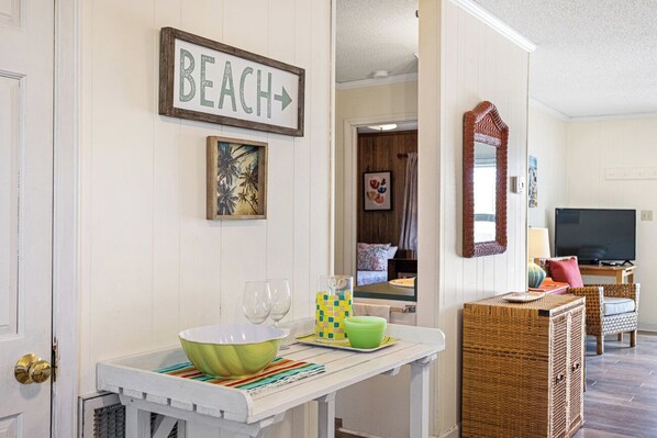 A room with a white wall, a framed 'BEACH' sign, a painting, a wicker side table with a bowl, glasses, and a checkered napkin holder. - Catch A Wave by Topsail Realty (Holly Ridge)