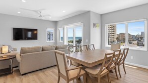 Una sala de estar moderna con sofá, mesa de comedor y sillas. Hay un televisor montado en la pared, y ventanas amplias con vistas a la ciudad.