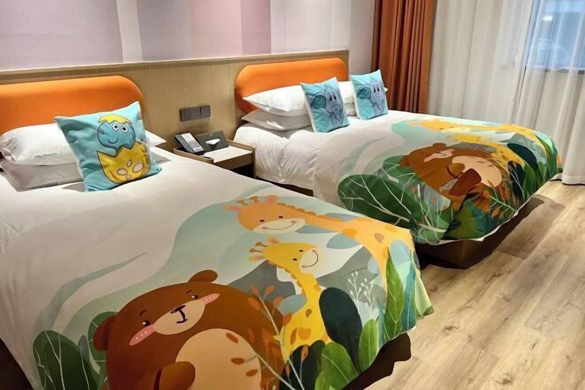 Two beds with colorful animal-themed bedding, a wooden nightstand with a phone, and a window with curtains.