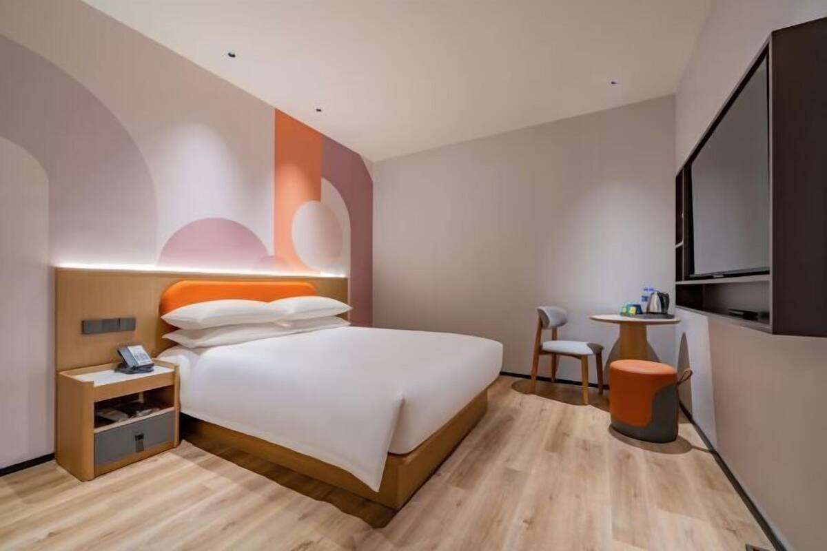 A modern hotel room with a bed, bedside table, chair, and a small table with a lamp.