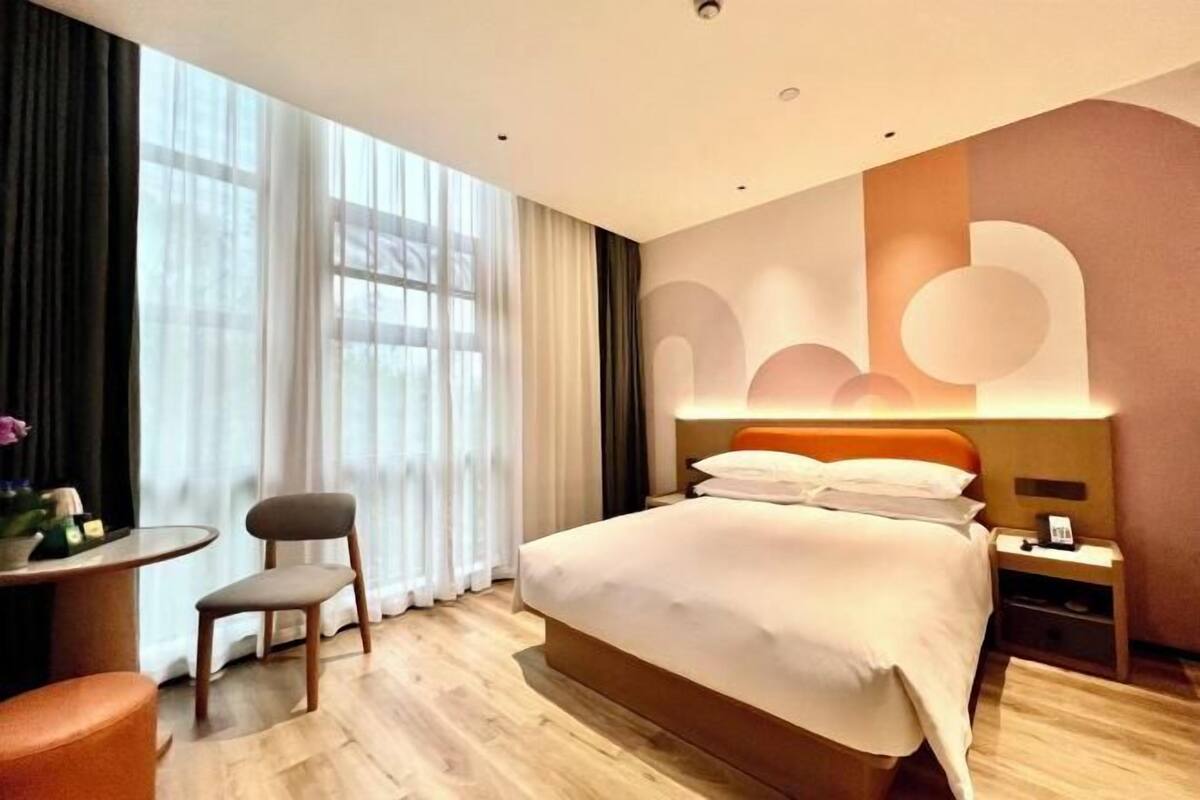 A modern hotel room with a large bed, a bedside table, a chair, and a view of the outdoors through large windows.