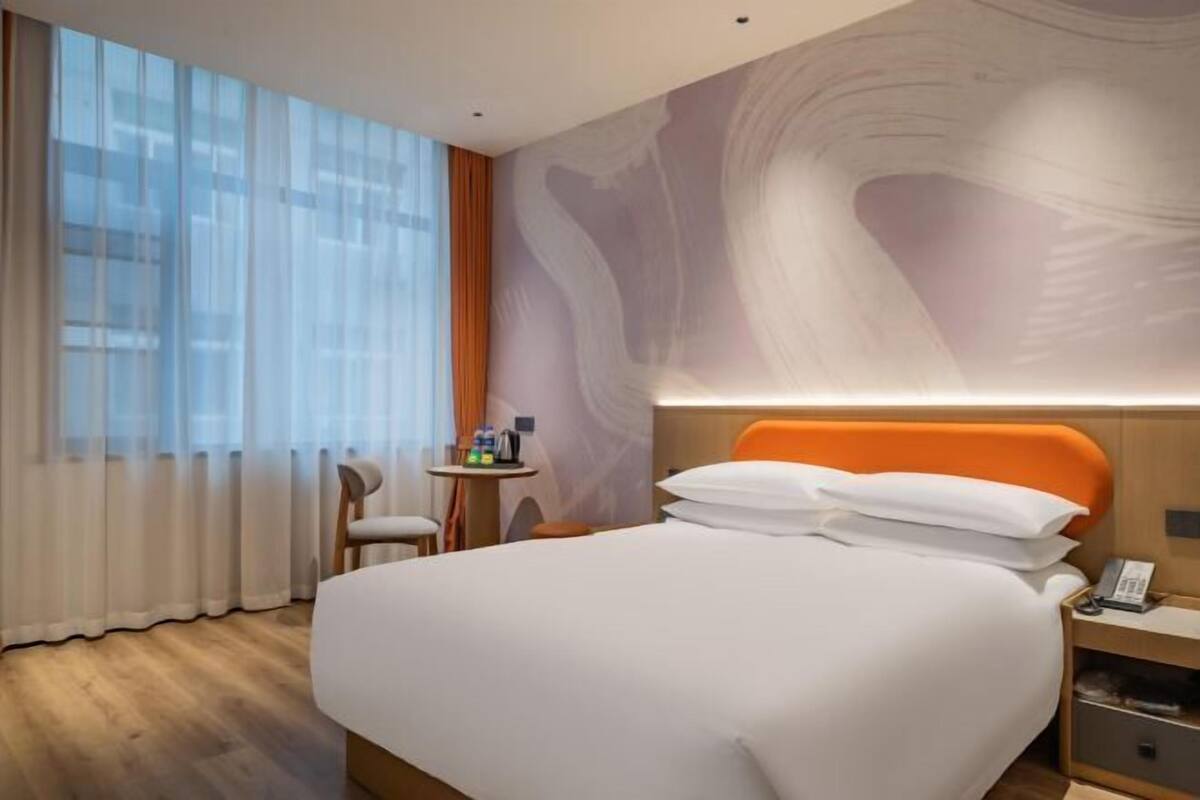 A hotel room with a bed, a nightstand, a small table with toiletries, a chair, and a large window with curtains.