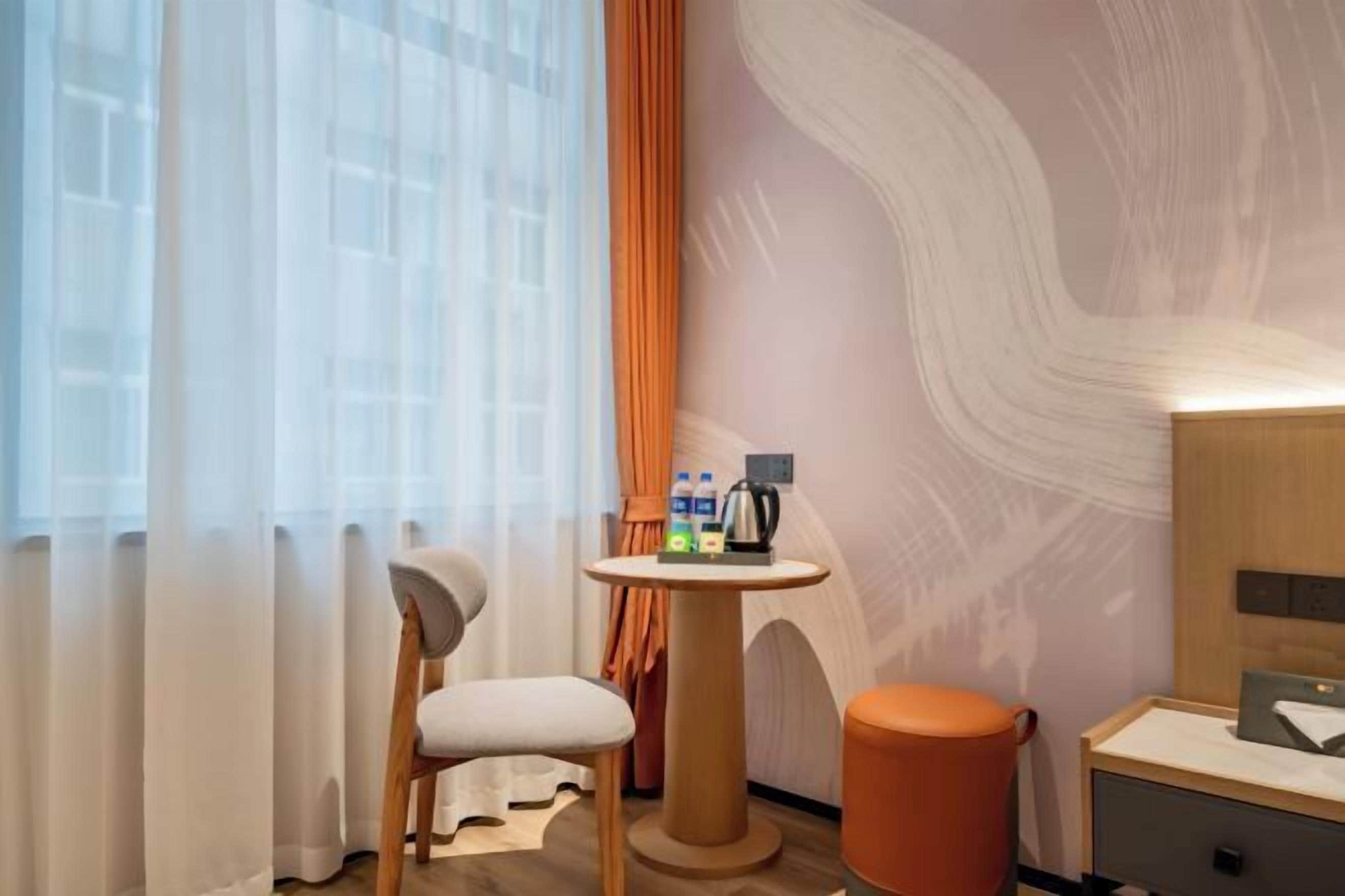 Une chambre d’hôtel avec une chaise en bois, une petite table ronde sur laquelle sont posés des articles de toilette, une lampe fixée au mur et une vue à travers des rideaux transparents.