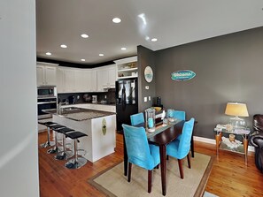 A modern kitchen with white cabinets, a black refrigerator, and a dining area with blue chairs. - Lucky Ducky by Carolina Retreats (Carolina Beach)