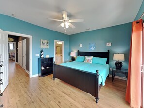 A bedroom with a bed, a nightstand, a dresser, a ceiling fan, and a door leading to another room. - Lucky Ducky by Carolina Retreats (Carolina Beach)