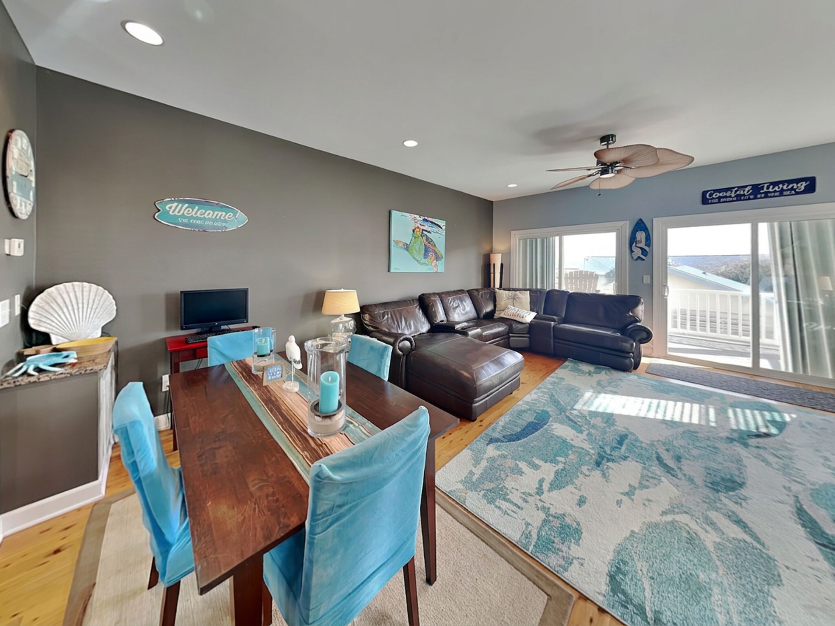 A modern living room with a dark leather sofa, a wooden dining table with blue chairs, a ceiling fan, and a large window with a view.