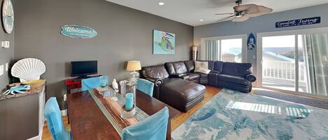 A modern living room with a dark leather sofa, a wooden dining table with blue chairs, a ceiling fan, and a large window with a view.