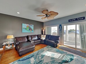 A living room with a brown leather sectional sofa, a ceiling fan, a colorful artwork, and a sign reading 'Coastal Living'. - Lucky Ducky by Carolina Retreats (Carolina Beach)