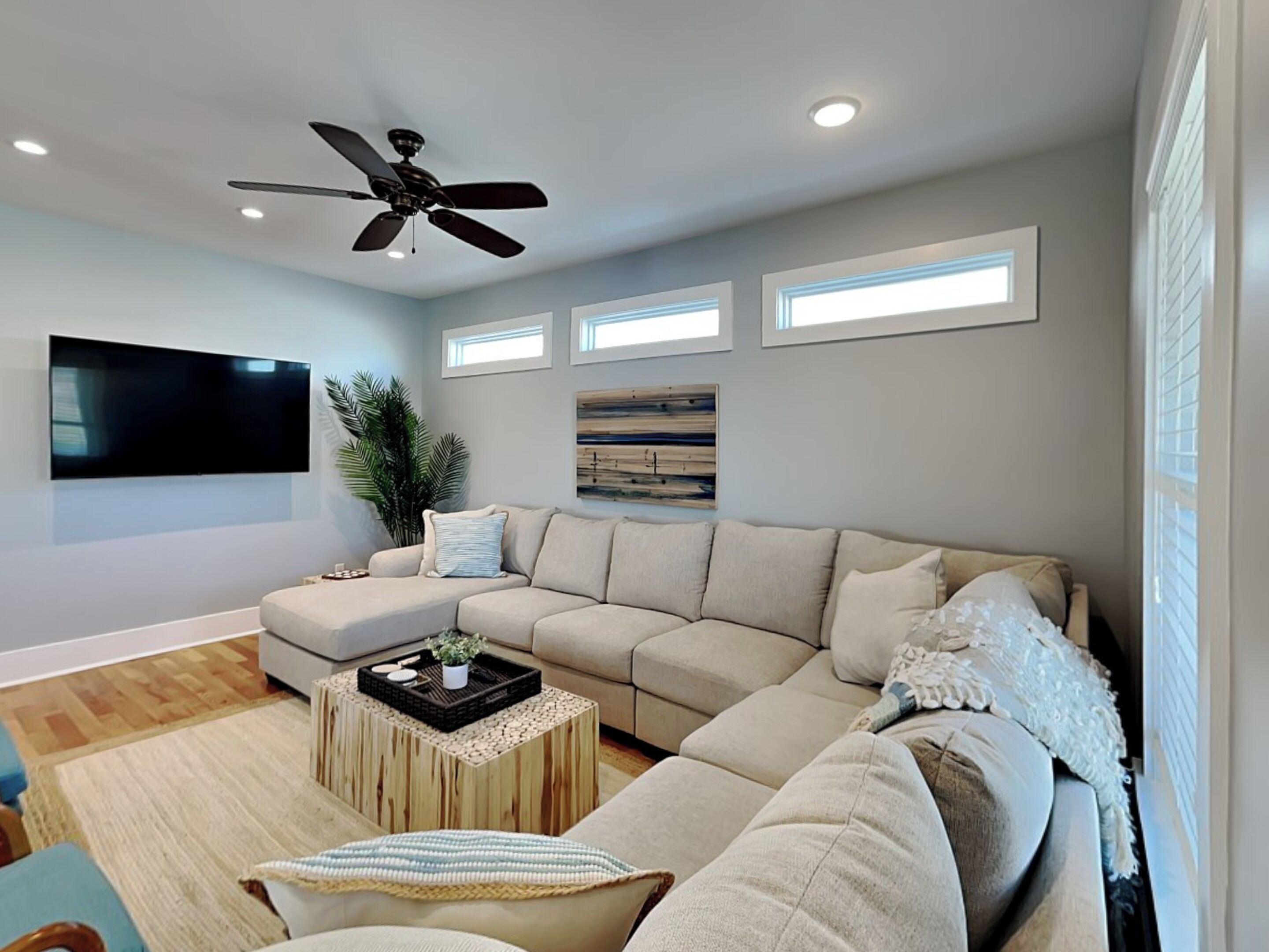 A modern living room with a sectional sofa, a ceiling fan, a flat-screen TV, and a large window with blinds.