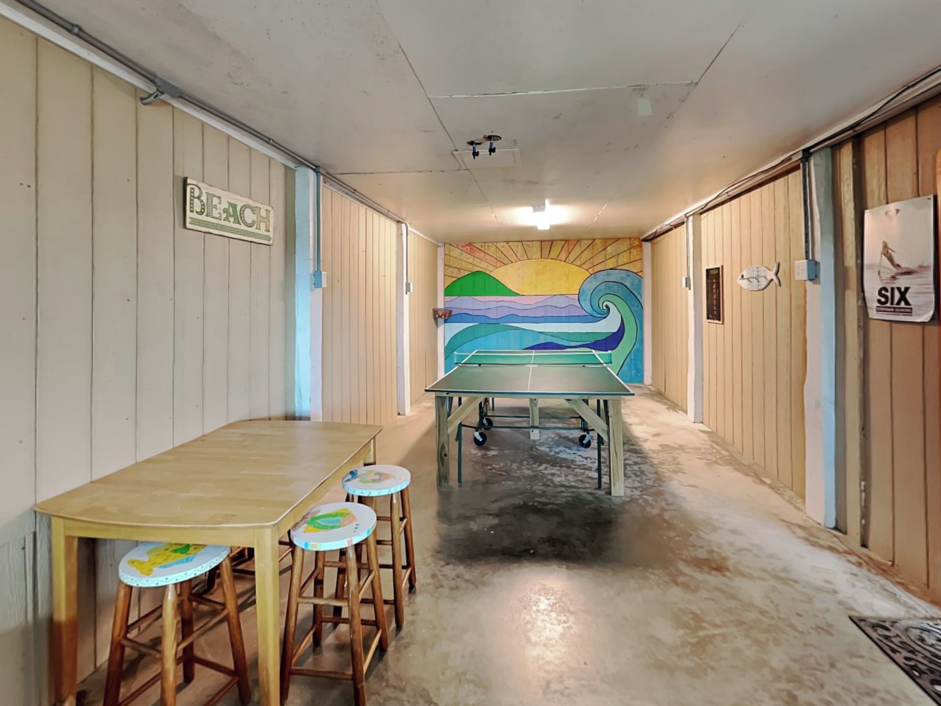 Un couloir avec une table de ping-pong, des tabourets en bois, une fresque murale et un panneau indiquant « PLAGE ».