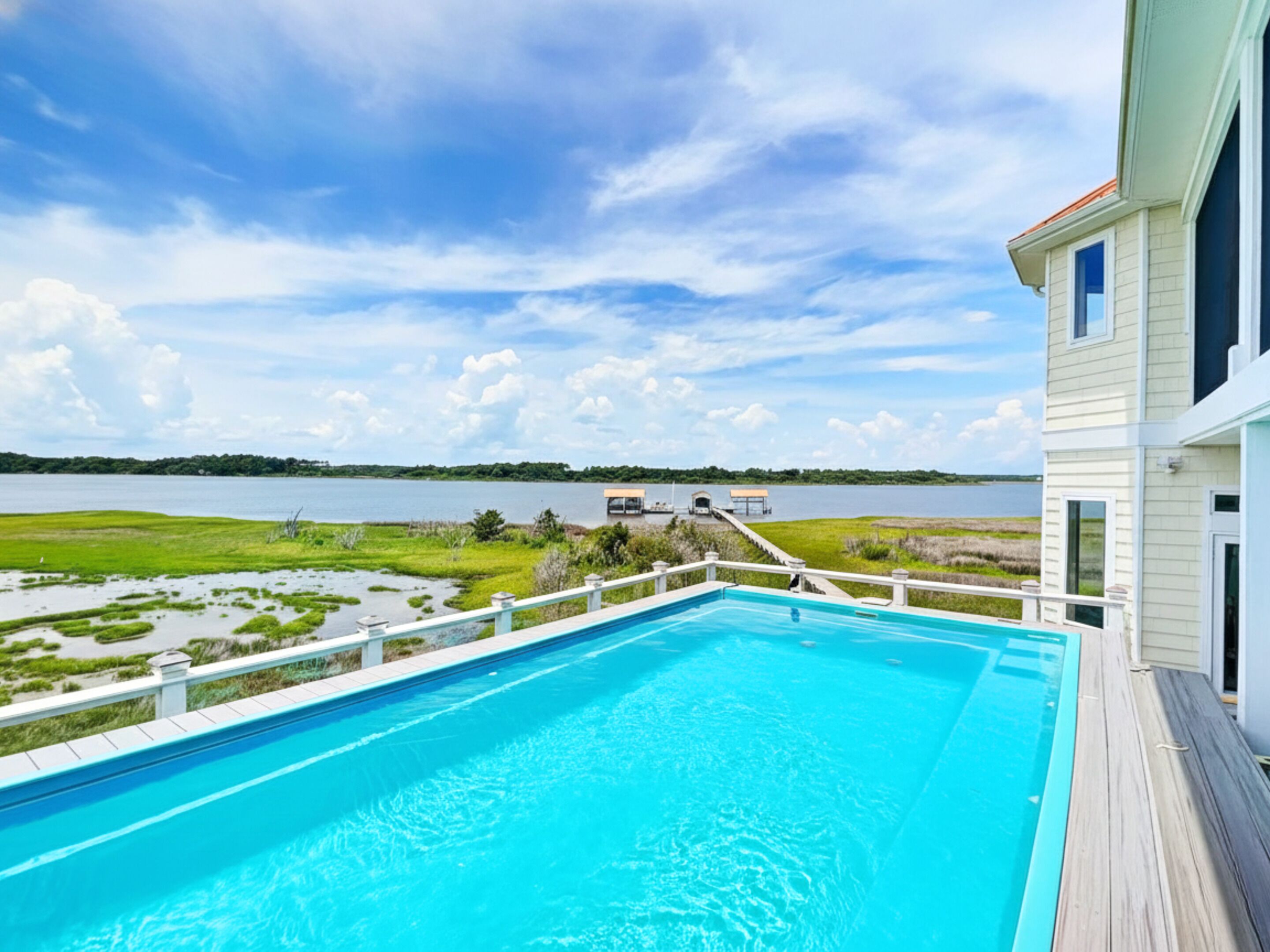 A swimming pool with a view of a lake and a dock extending into the water, surrounded by a deck and a building with a white exterior.
